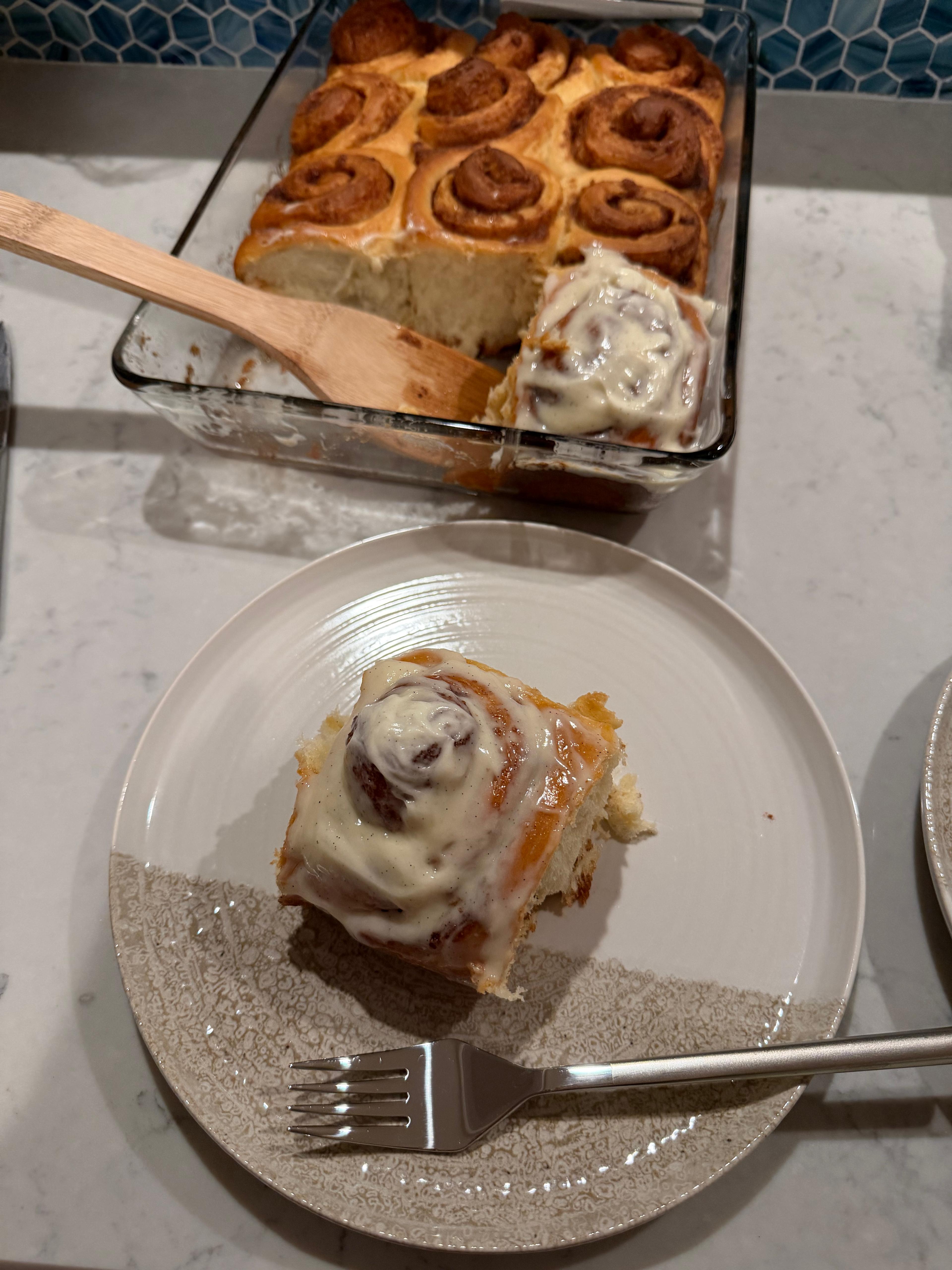 A frosted cinnamon roll on a plate with a tray of cinnamon rolls in the background.