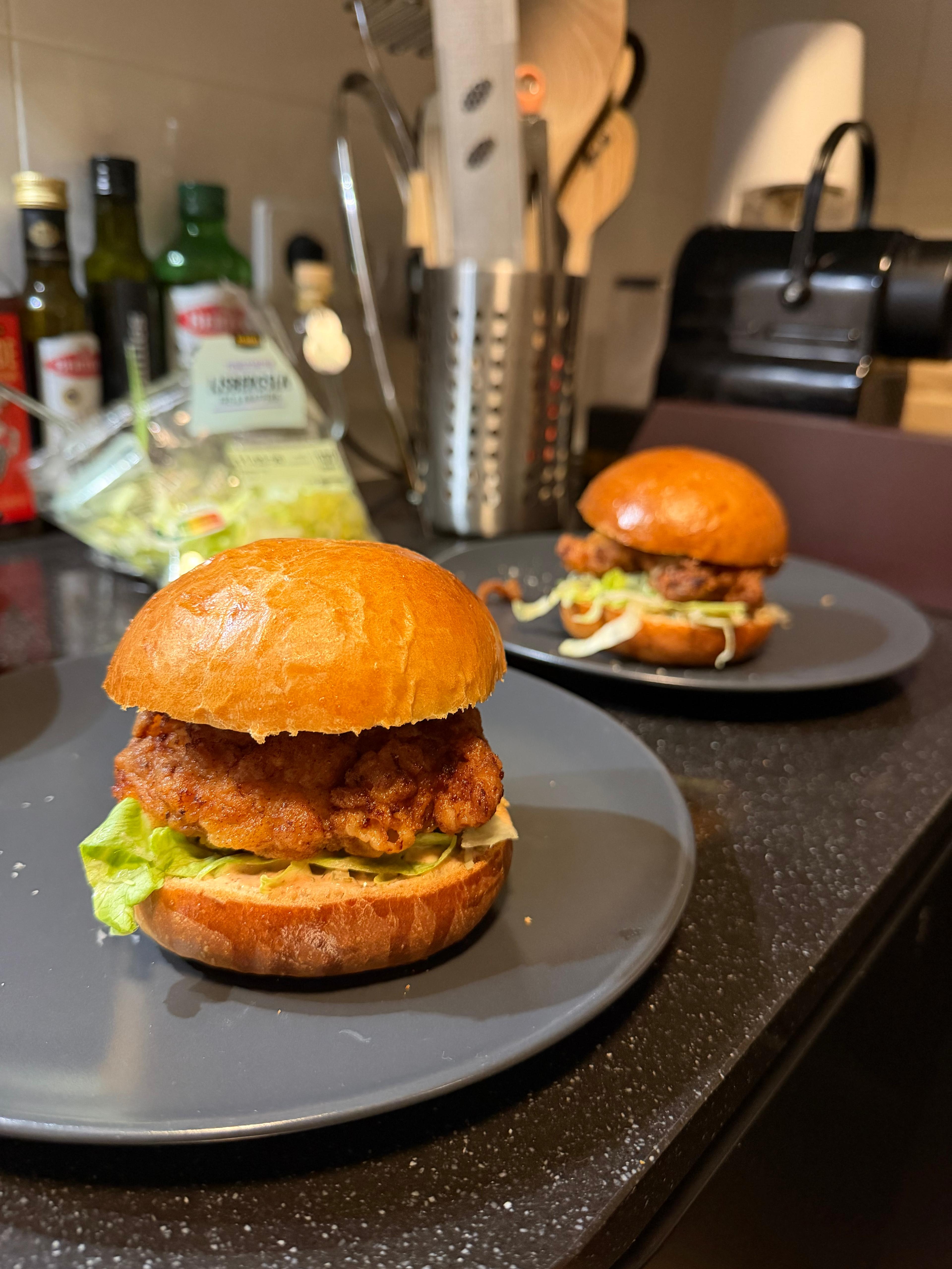 Two milk buns sitting on dark grey plates, in between a salad leaf and a piece of fried chicken.