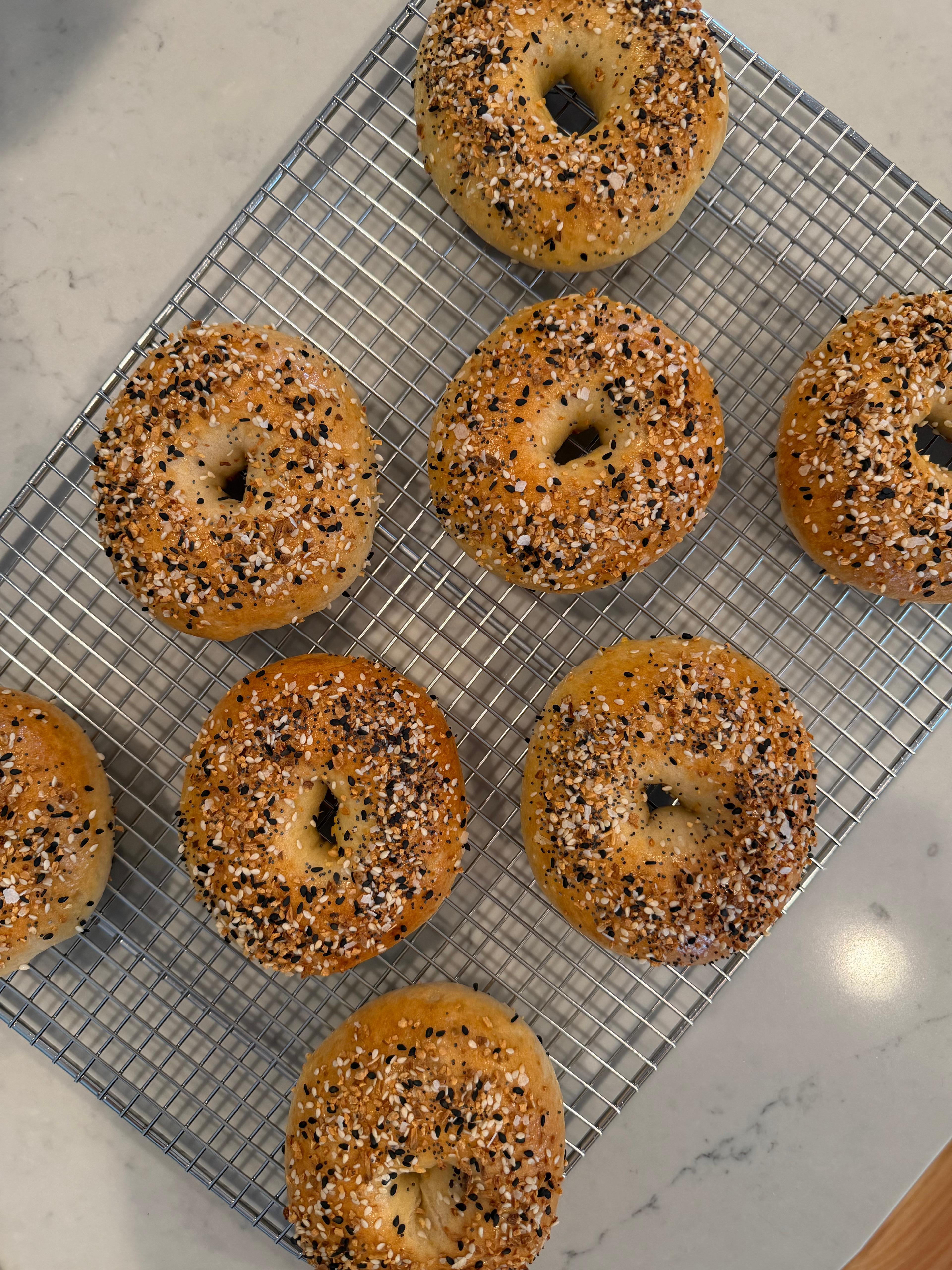 Whole everything bagels on a cooling rack, on a white marble kitchen counter.